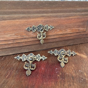 May include: Three antique brass drawer pulls with ornate floral and scroll designs. The pulls are mounted on a wooden surface, showcasing their intricate details and aged patina. The hardware is a decorative element for furniture.