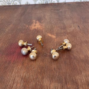 May include: A collection of vintage brass cabinet knobs with a rounded, ball-like design. The knobs are a shiny gold color and are scattered on a dark wooden surface. The hardware is likely for furniture restoration or DIY projects.