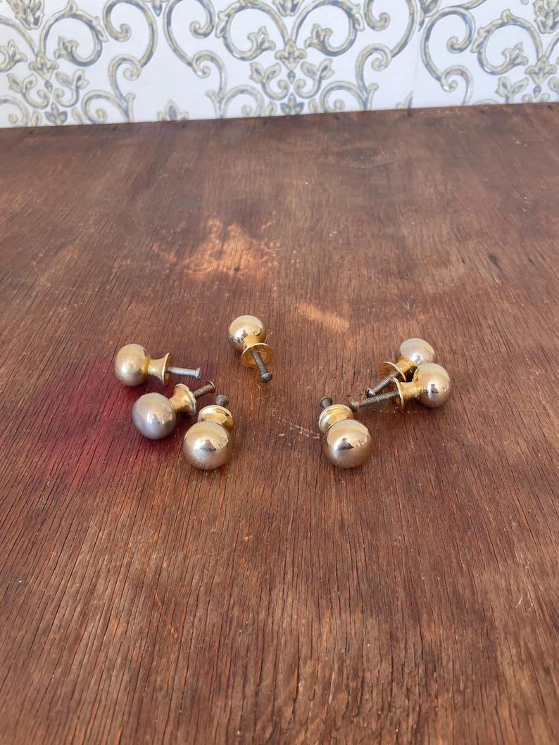 May include: A collection of vintage brass cabinet knobs with a rounded, ball-like design. The knobs are a shiny gold color and are scattered on a dark wooden surface. The hardware is likely for furniture restoration or DIY projects.