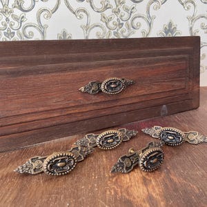 May include: A set of four antique-style brass drawer pulls featuring an oval center with a floral motif and elongated, decorative pointed ends. One pull is attached to a dark brown wooden drawer front, while three others are arranged in front.