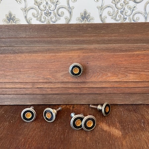 Könnte beinhalten: Eine Sammlung von Vintage-Schrankknöpfen mit silbernem Finish, schwarzem Rand und einem orange-gelben Mittelkreis. Die Knöpfe werden auf einer Holzoberfläche präsentiert, einer ist an einer Holzschublade befestigt.