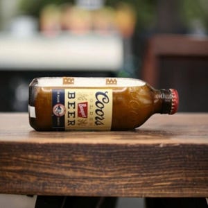 May include: A brown glass Coors Banquet beer bottle laid on its side, featuring a cream-colored label with blue and red text. The bottle has a red cap and a rectangular section cut out from the top, revealing a white interior surface.