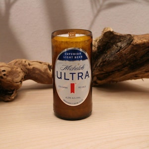 May include: A candle housed in a repurposed amber glass Michelob Ultra beer bottle. The label features the text Superior Light Beer, Michelob Ultra, 2.6 Carbs, 95 Cals, and 4.2% Alc./Vol. A wooden wick is visible at the top of the white wax.