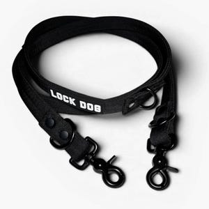 Może przedstawiać: Czarna smycz dla psa z prostokątną naszywką z białym napisem LOCK DOG. Smycz wykonana jest z wytrzymałej taśmy i zawiera dwa czarne metalowe obrotowe karabińczyki, dwa metalowe pierścienie typu D oraz wzmocnione szwy w punktach łączenia.