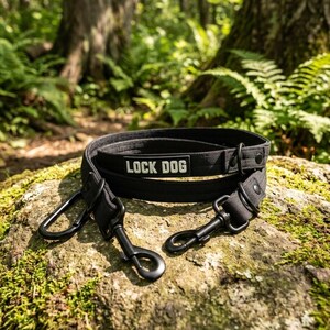 Może przedstawiać: Czarna smycz dla psa wykonana z wytrzymałej tkaniny z prostokątną naszywką z białym napisem LOCK DOG. Smycz posiada dwa czarne metalowe karabińczyki obrotowe, kilka metalowych kółek w kształcie litery D oraz czarne nity mocujące taśmę.