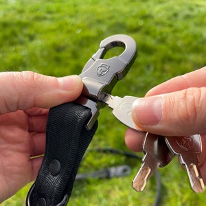 Può includere: Un moschettone in metallo nero con un cinturino in nylon nero attaccato. Il moschettone ha un logo argentato ed è aperto per rivelare una chiave argentata con un logo. La chiave è tenuta da una mano.