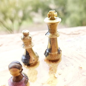 May include: Three wooden chess pieces, a king, a bishop, and a pawn, are arranged on a wooden surface. The king is in the center, the bishop is to the left, and the pawn is to the right. The pieces are painted in a gold and black color scheme.
