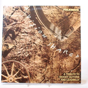 May include: A vinyl record album cover featuring a brown and tan textured background with a white wheel and the text "A Vision Shared" and "A Tribute to Woody Guthrie and Leadbelly".