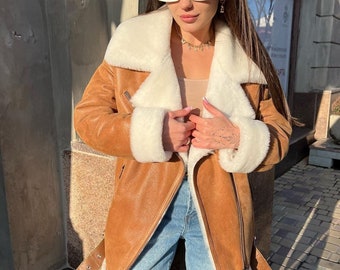 May include: A tan jacket with a white faux fur lining, wide lapels, and cuffs. The jacket features a diagonal front zipper, side pockets with zippers, and a buckled belt at the hem. It is worn over a beige top and light blue denim jeans.