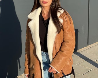 May include: A brown shearling jacket with a cream-colored shearling lining and collar. The jacket has a belted waist, black trim, and a classic aviator style. The model is wearing sunglasses and blue jeans.