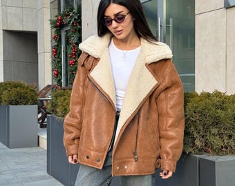 May include: A brown faux leather jacket with a cream-colored sherpa lining, a wide notched collar, and a diagonal zipper closure. The jacket features long sleeves with buckled cuffs, a white crew neck t-shirt, and blue denim jeans.
