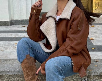 May include: A brown jacket with a cream-colored faux shearling lining and collar, worn with blue denim jeans and brown suede cowboy boots featuring decorative stitching. The jacket has a front zipper, long sleeves with buttoned cuffs, and a side belt.