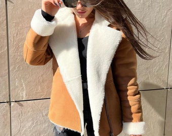 May include: A tan suede jacket with a white faux shearling lining, wide lapels, and long sleeves with white cuffs. The jacket features a silver-tone zipper closure and is worn over a black top and distressed black denim jeans.