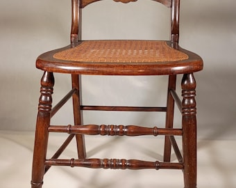 May include: A dark brown wooden chair featuring a woven cane seat, a curved backrest with two horizontal slats, and turned legs connected by horizontal stretchers. The wood has a polished finish, and the seat has a natural tan color.