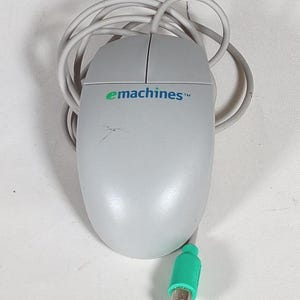 Puede incluir: Un ratón de ordenador gris claro con dos botones y una rueda de desplazamiento. La superficie superior presenta el logotipo de eMachines en verde y azul. Un cable gris enrollado está unido, terminando en un conector PS/2 verde redondo.