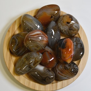 May include: A collection of agate stones in various shades of brown, grey, and orange. The stones are smooth and polished, with intricate patterns of banding and swirls. The stones are displayed on a wooden plate.