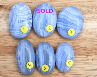 May include: Six oval-shaped blue lace agate stones with white banding patterns. Each stone is marked with a yellow circular sticker numbered one through six. The second stone has the word SOLD printed in bright pink text across its surface.