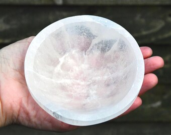 May include: A circular bowl carved from translucent white selenite crystal. The bowl features a smooth, polished rim and a textured, crystalline interior with natural mineral inclusions and visible fractures throughout the stone structure.
