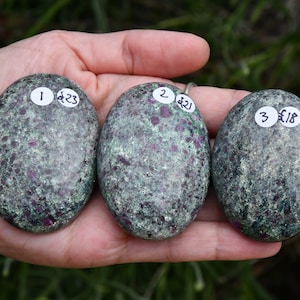 May include: Three oval-shaped green and pink stones with red flecks. Each stone has a white sticker with a number and price in pounds sterling. The stones are labelled 1 £23, 2 £21, and 3 £18.
