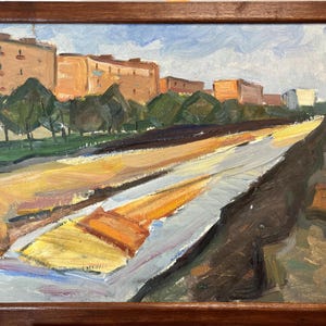 May include: An oil painting in a brown wooden frame depicting an urban landscape with orange apartment buildings, green trees, and a wide, light-coloured road or path stretching into the distance under a pale blue sky with white clouds.
