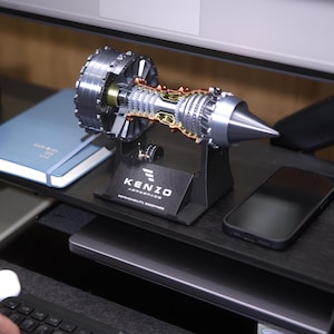 May include: A silver and black model of a jet engine on a black stand with the text "KENZO AEROSPACE DEPENDABILITY REDEFINED".