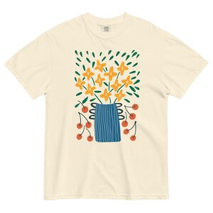 Op de afbeelding: Crèmekleurig T-shirt met een bloemenontwerp. De afbeelding bevat oranje bloemen in een blauwe vaas, groene bladeren en rode kersen. Het ontwerp is een grillige, artistieke illustratie.