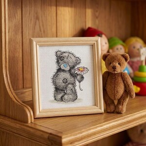 May include: A square wooden frame containing a cross-stitch embroidery of a grey teddy bear with a blue nose holding a white and pink daisy. Next to the frame stands a small, fuzzy brown teddy bear on a wooden shelf with other toys.