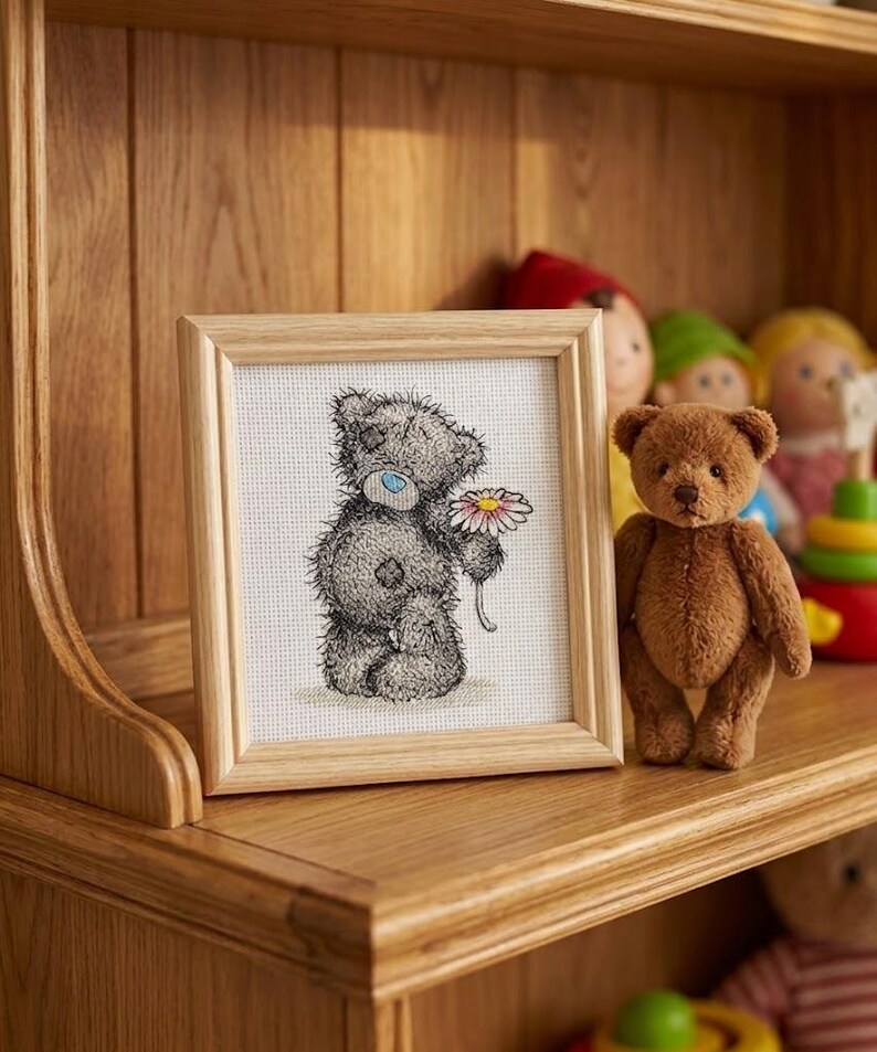 May include: A square wooden frame containing a cross-stitch embroidery of a grey teddy bear with a blue nose holding a white and pink daisy. Next to the frame stands a small, fuzzy brown teddy bear on a wooden shelf with other toys.