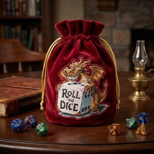 May include: A deep red velvet drawstring pouch featuring an embroidered dragon holding a large white die with the text Roll the Dice. The bag has gold-colored cords and is surrounded by several multi-sided gaming dice on a dark wooden surface.