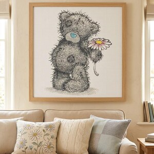 May include: A square framed cross-stitch artwork featuring a shaggy gray teddy bear with a blue nose and a patch on its eye, holding a single daisy with white petals and a yellow center, set against a white grid fabric background.