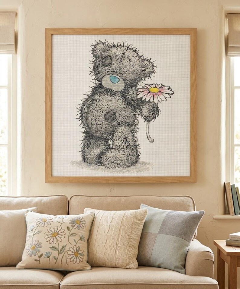 May include: A square framed cross-stitch artwork featuring a shaggy gray teddy bear with a blue nose and a patch on its eye, holding a single daisy with white petals and a yellow center, set against a white grid fabric background.