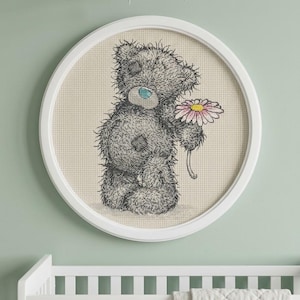 May include: A circular framed cross-stitch embroidery featuring a grey teddy bear with a blue nose and patches, holding a pink daisy with a yellow center. The bear is rendered in a sketch-like style on a beige grid fabric, enclosed in a white frame.