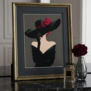 May include: A framed cross-stitch embroidery depicting a figure in a wide-brimmed black hat adorned with red flowers, wearing a black off-the-shoulder garment and black gloves, set against a dark gray background within a gold-toned frame.
