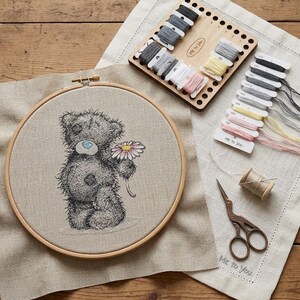 May include: A circular wooden embroidery hoop containing a beige fabric panel with a stitched grey teddy bear holding a pink and yellow flower. Includes a wooden thread organizer, assorted embroidery floss, a spool of thread, and metal scissors.