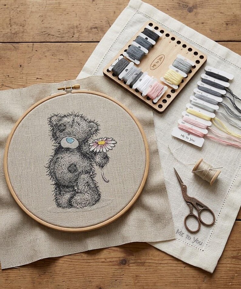 May include: A circular wooden embroidery hoop containing a beige fabric panel with a stitched grey teddy bear holding a pink and yellow flower. Includes a wooden thread organizer, assorted embroidery floss, a spool of thread, and metal scissors.