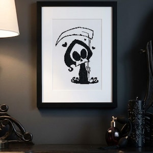 May include: A framed cross-stitch embroidery featuring a black and white pixelated illustration of a grim reaper character holding a scythe, accented with two small hearts on either side, enclosed in a simple black frame with a white mat board.