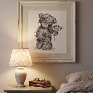 May include: A framed square wall art piece featuring a sketch of a grey teddy bear with a blue nose holding a pink and white daisy. The artwork is encased in a distressed off-white wooden frame with a textured matting, displayed on a wall.