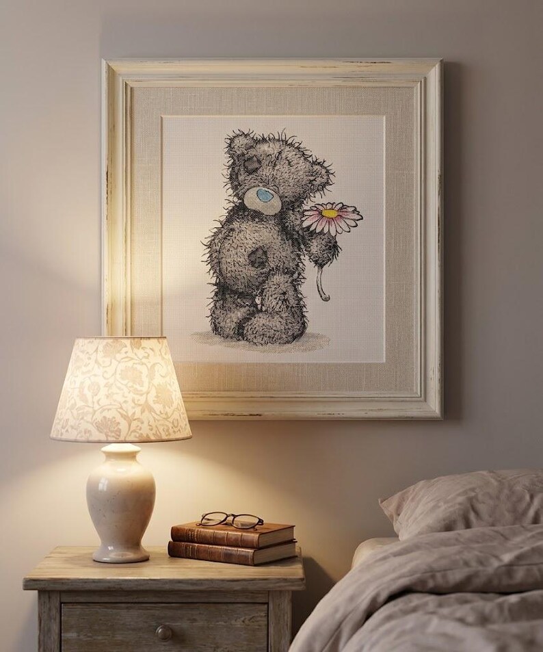 May include: A framed square wall art piece featuring a sketch of a grey teddy bear with a blue nose holding a pink and white daisy. The artwork is encased in a distressed off-white wooden frame with a textured matting, displayed on a wall.