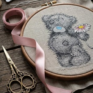 May include: A round wooden embroidery hoop containing a beige linen fabric with a stitched gray teddy bear holding a pink and white daisy. A light pink ribbon, a pair of ornate brass scissors, and a spool of thread are arranged around the hoop.
