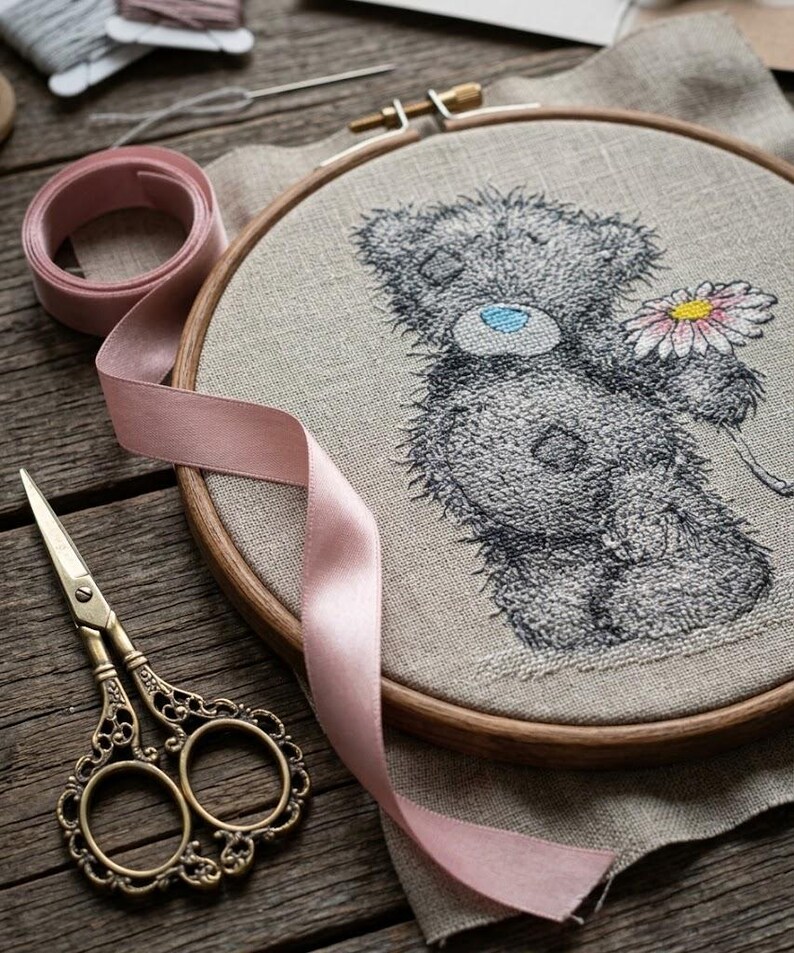 May include: A round wooden embroidery hoop containing a beige linen fabric with a stitched gray teddy bear holding a pink and white daisy. A light pink ribbon, a pair of ornate brass scissors, and a spool of thread are arranged around the hoop.