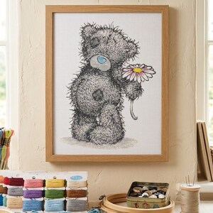 May include: A framed cross-stitch embroidery featuring a shaggy gray teddy bear with a blue nose and a patch on its chest, holding a single daisy with white petals and a yellow center. The artwork is displayed in a light-toned wooden frame.
