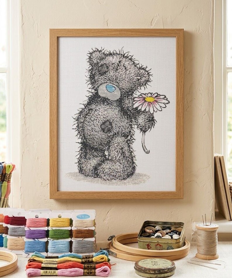 May include: A framed cross-stitch embroidery featuring a shaggy gray teddy bear with a blue nose and a patch on its chest, holding a single daisy with white petals and a yellow center. The artwork is displayed in a light-toned wooden frame.