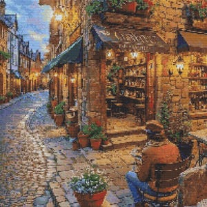 Op de afbeelding: Een smalle straat met keien in een Europees dorp met een café aan de rechterkant. Het café heeft een groene luifel en een bord met de tekst "Café". Er staan potplanten en bloemen langs de straat.