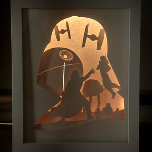 Puede incluir: Un marco blanco con un recorte de papel marrón claro de Darth Vader, una Estrella de la Muerte y cazas TIE. El papel está iluminado desde atrás, creando un efecto de silueta.