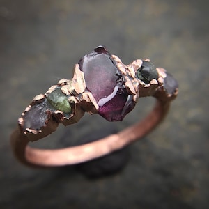 Peut inclure: Une bague artisanale avec un anneau en cuivre et un groupe de pierres précieuses brutes. La bague présente une grosse pierre centrale violet foncé, flanquée de pierres vertes et violettes plus petites. Le sertissage a un design texturé et organique.