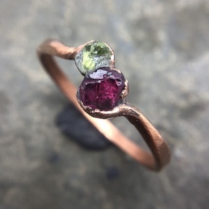 May include: A ring featuring a thin, textured copper-toned band set with two raw, uncut gemstones. One stone is a translucent light green, while the other is a deep, saturated magenta-red. The stones are held in place by metal prongs.