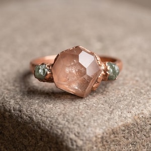 Peut inclure: Une bague aux tons cuivrés ornée d'une grande pierre centrale translucide couleur pêche à facettes, entourée de deux petites pierres brutes vert sauge. L'anneau métallique présente un aspect texturé et organique avec des griffes.