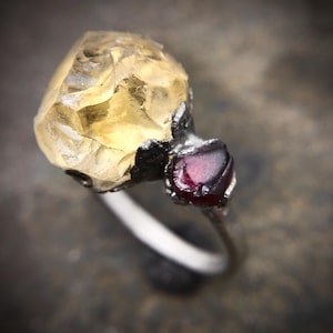 Peut inclure: Une bague en argent ornée d'une grande pierre précieuse jaune pâle et facettée et d'une plus petite pierre précieuse rouge foncé. La bague a un design organique unique, avec les pierres serties dans un sertissage en métal foncé.