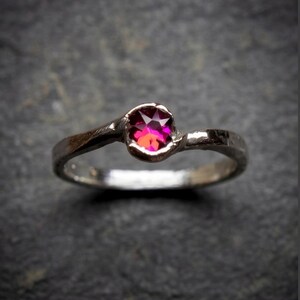 May include: A silver-toned ring featuring a single, round, faceted, vibrant magenta-pink gemstone set in a smooth, organic, slightly asymmetrical band with a polished metallic finish that reflects light across its curved, textured surface.