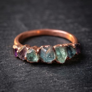 Peut inclure: Une bague en cuivre ornée d'une rangée de pierres précieuses brutes et non taillées dans des tons de vert et de violet. L'anneau est en cuivre poli et les pierres sont serties dans une monture métallique texturée aux formes irrégulières.